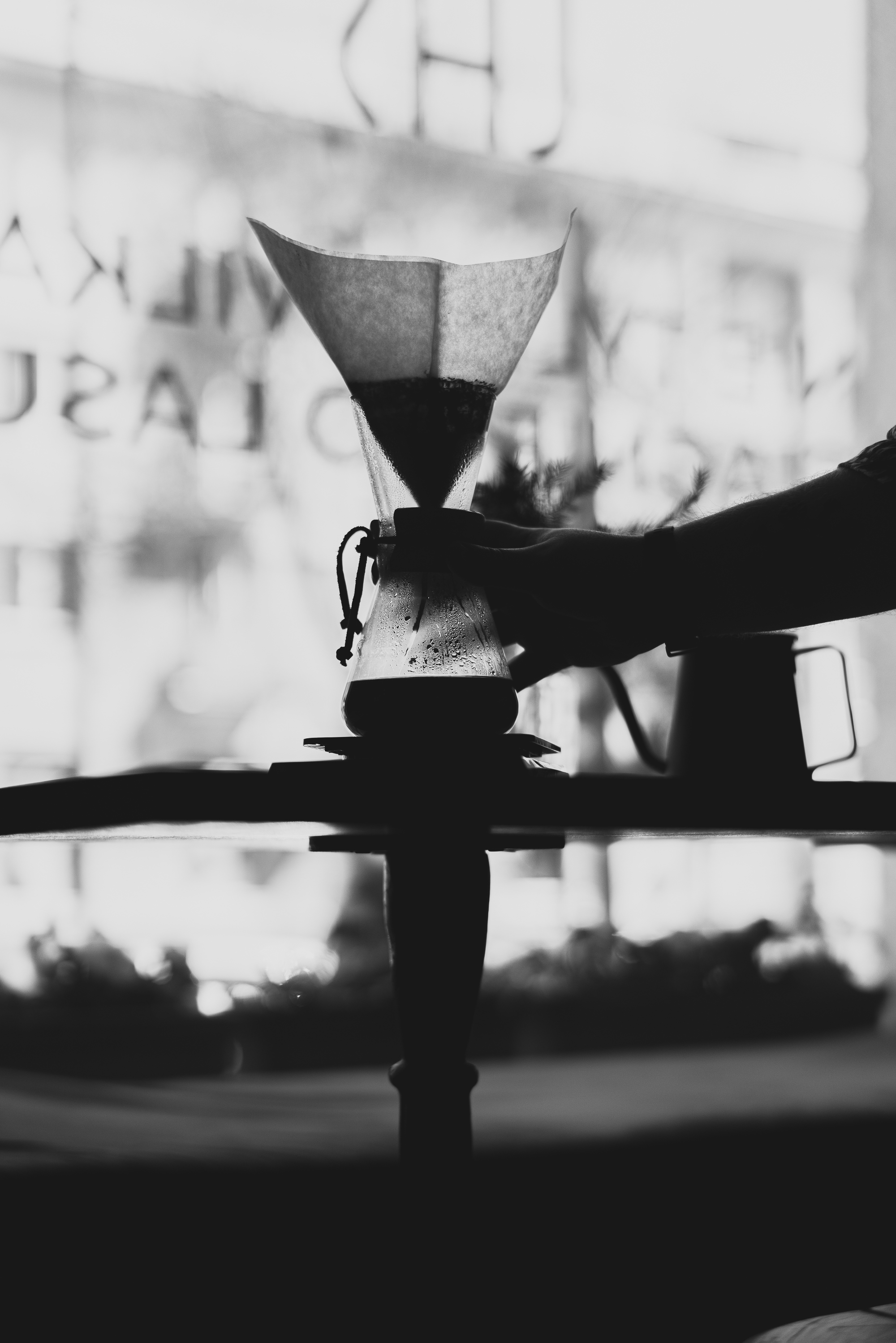 Hand holding a Chemex coffee maker with a paper filter on a table, silhouetted against a bright window with blurred text in the background, creating a calm and contemplative atmosphere